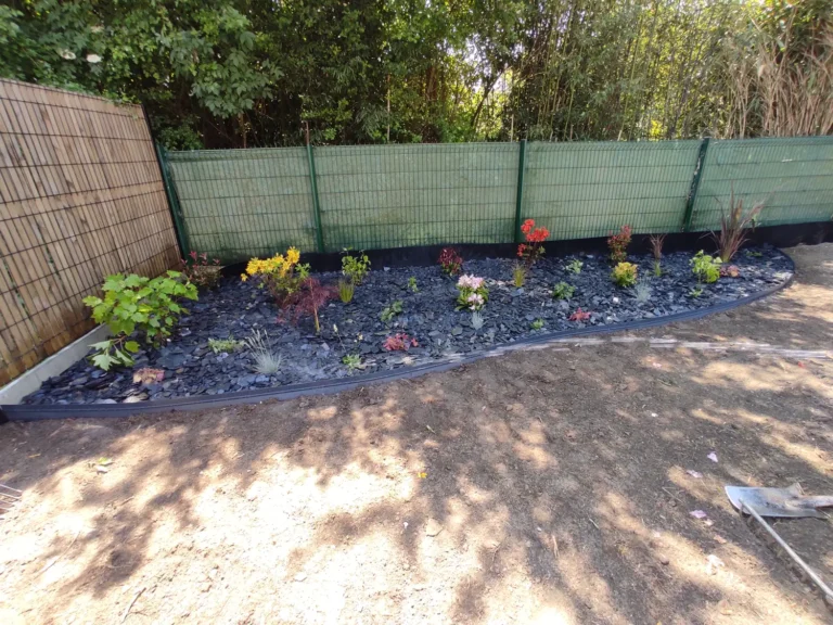 Parterre fleuri avec paillis bleu contre une clôture verte, entouré de béton et de sable